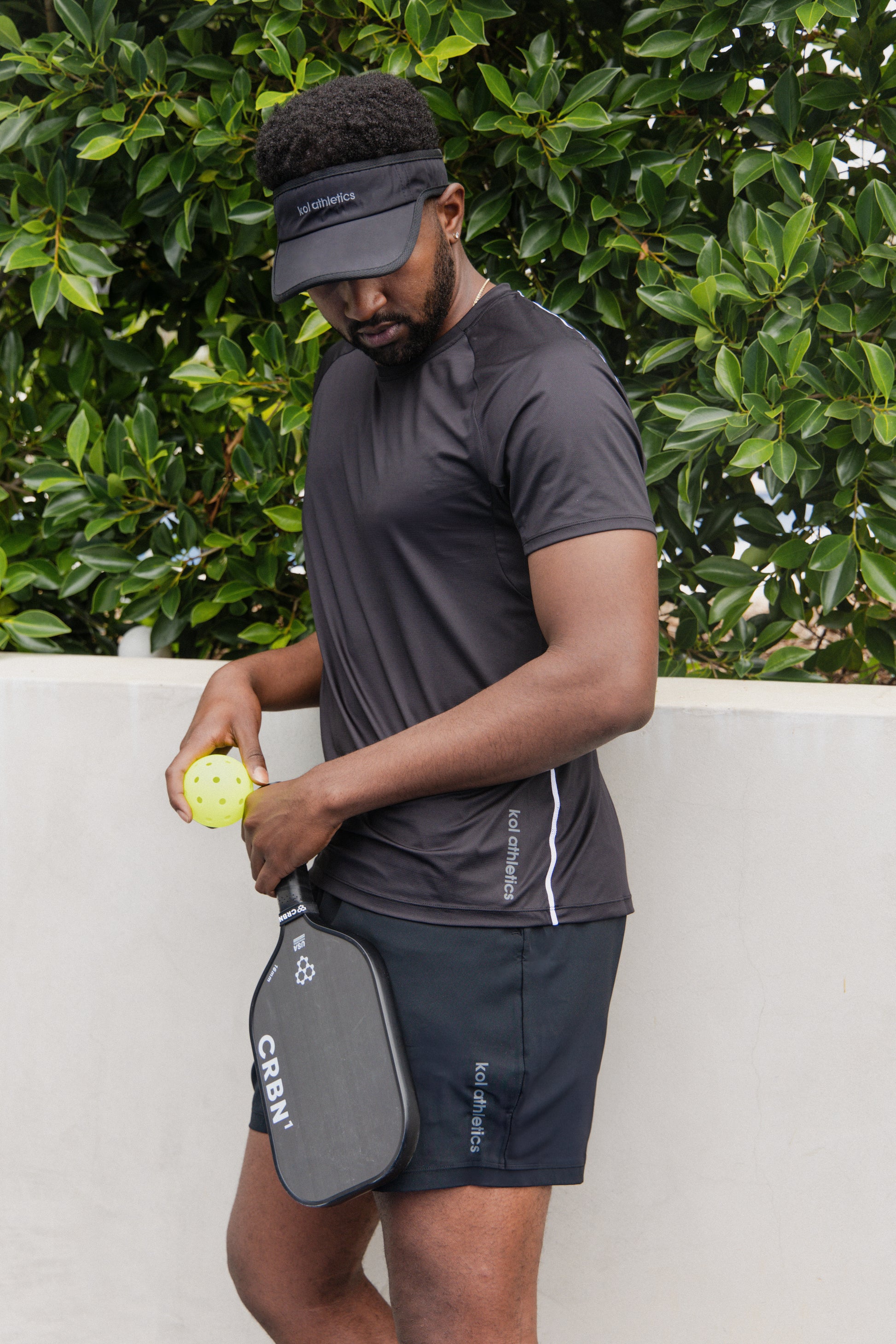 male pickleball player wearing black unlined athletic shorts and a black athletic shirt and a black visor, holding a pickleball and a pickleball paddle